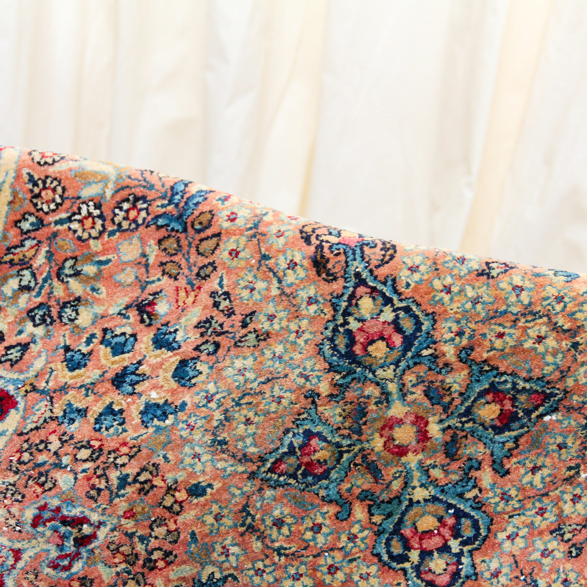 A close-up of a patterned area rug with a distressed traditional design in peach, blue, red, and beige tones, draped against a soft cream fabric backdrop.