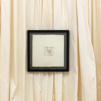 A square black-framed art print with a white mat and a small central sketch, displayed against a backdrop of draped cream-colored fabric.