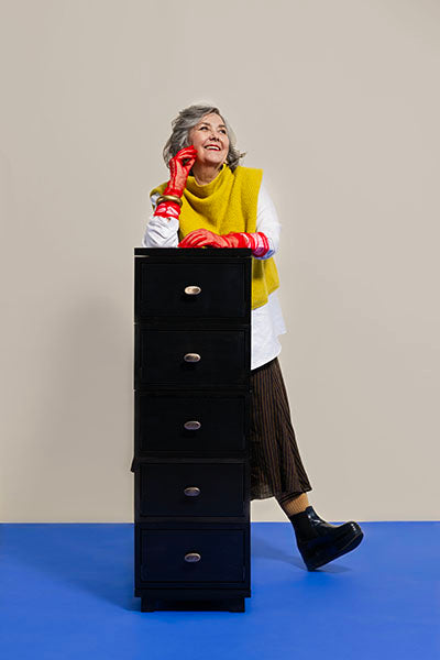 Woman wearing red gloves and a yellow vest standing next to a black dresser.