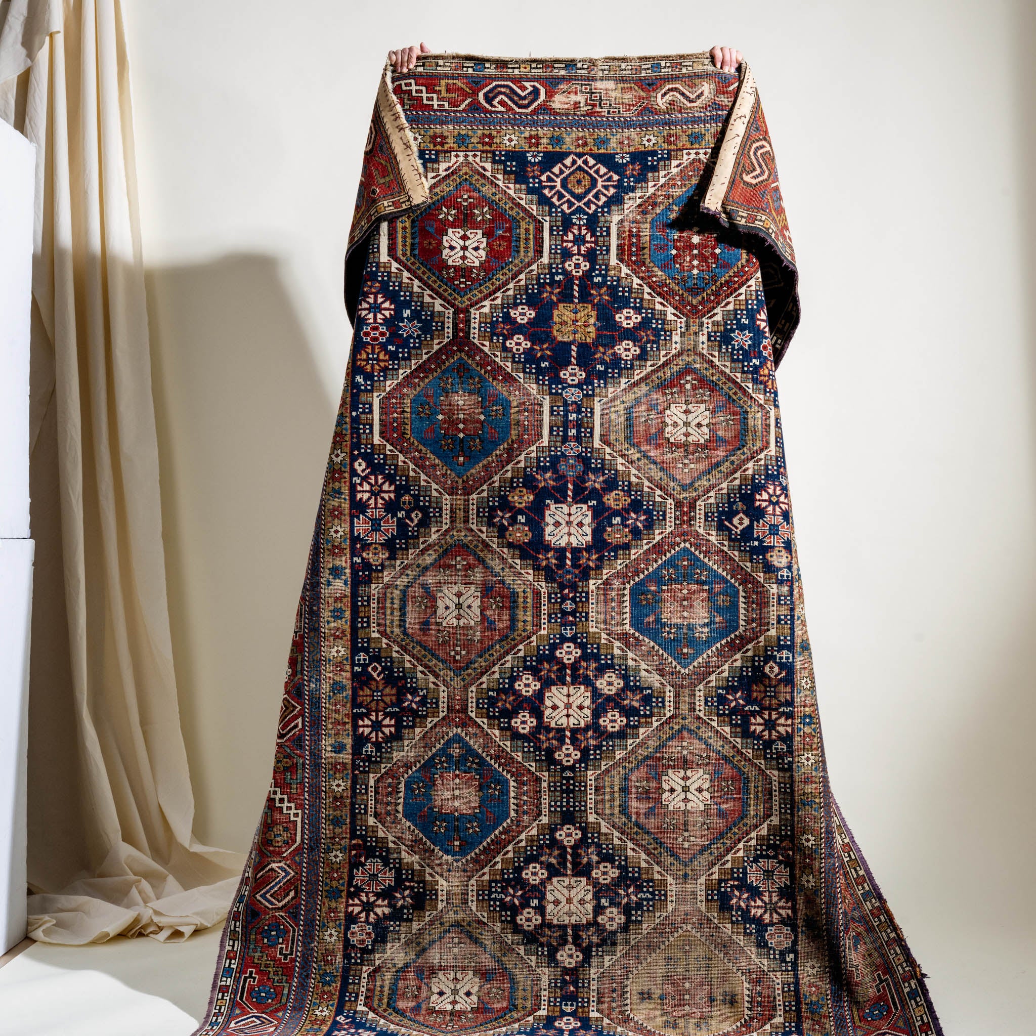 Decorative rug with intricate patterns draped held up by a person against a white wall