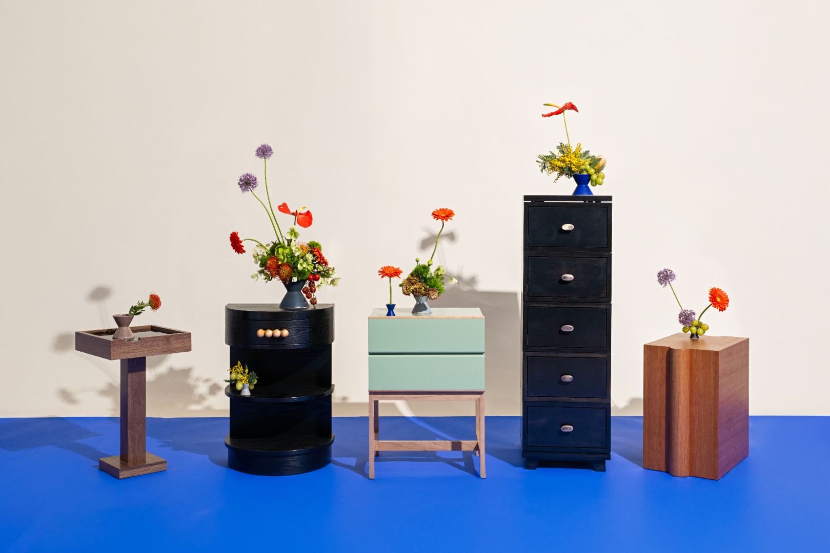 Collection of handcrafted furniture pieces with floral arrangements on a blue floor and white wall background