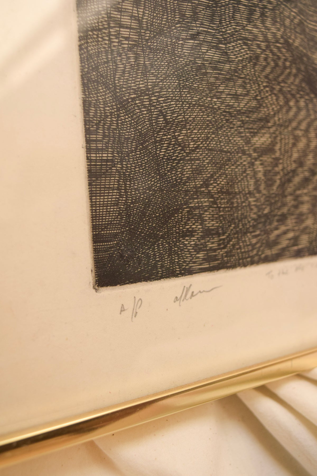 A close-up of a framed art print featuring a black and gray textured etching on warm off-white paper, with a gold frame edge visible at the bottom.