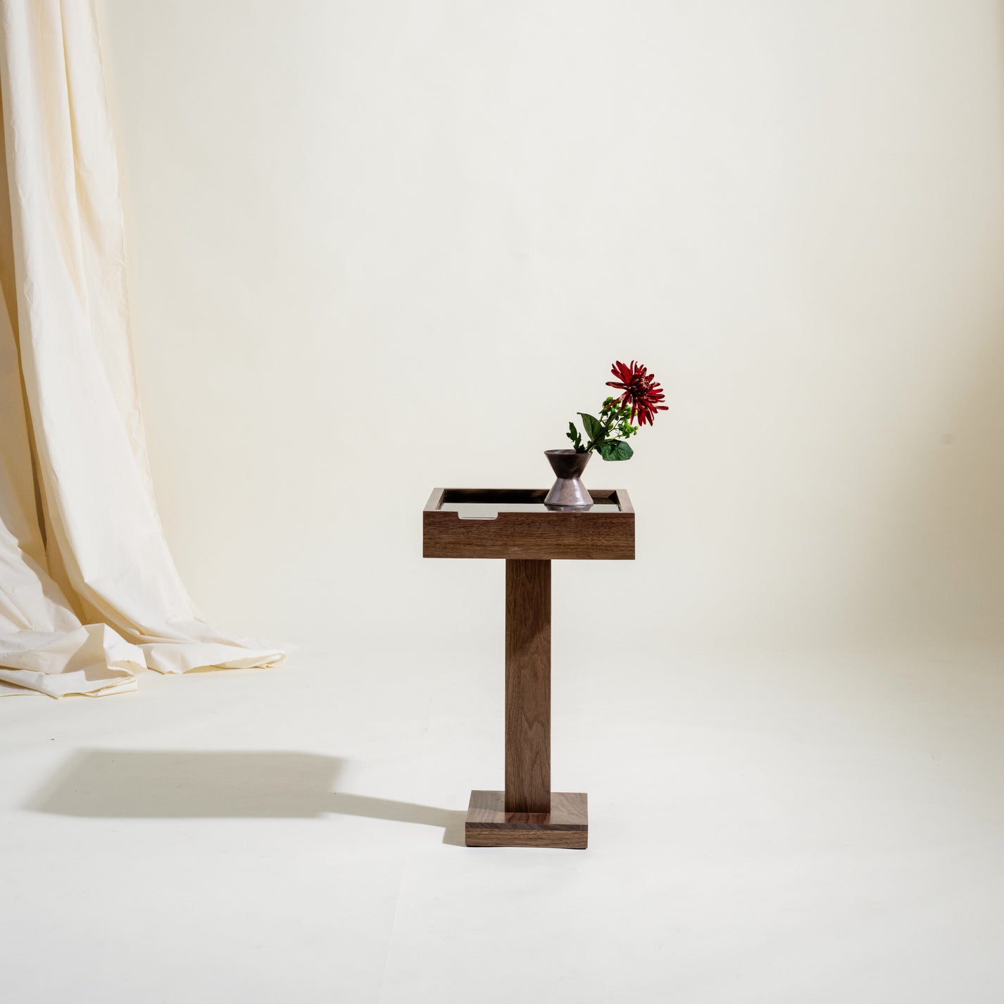 Handcrafted wooden side table with a vase of flowers on a white background. Table has a small square base on the floor and a square column coming up from the base with a rectangular top that is larger is area than the base.