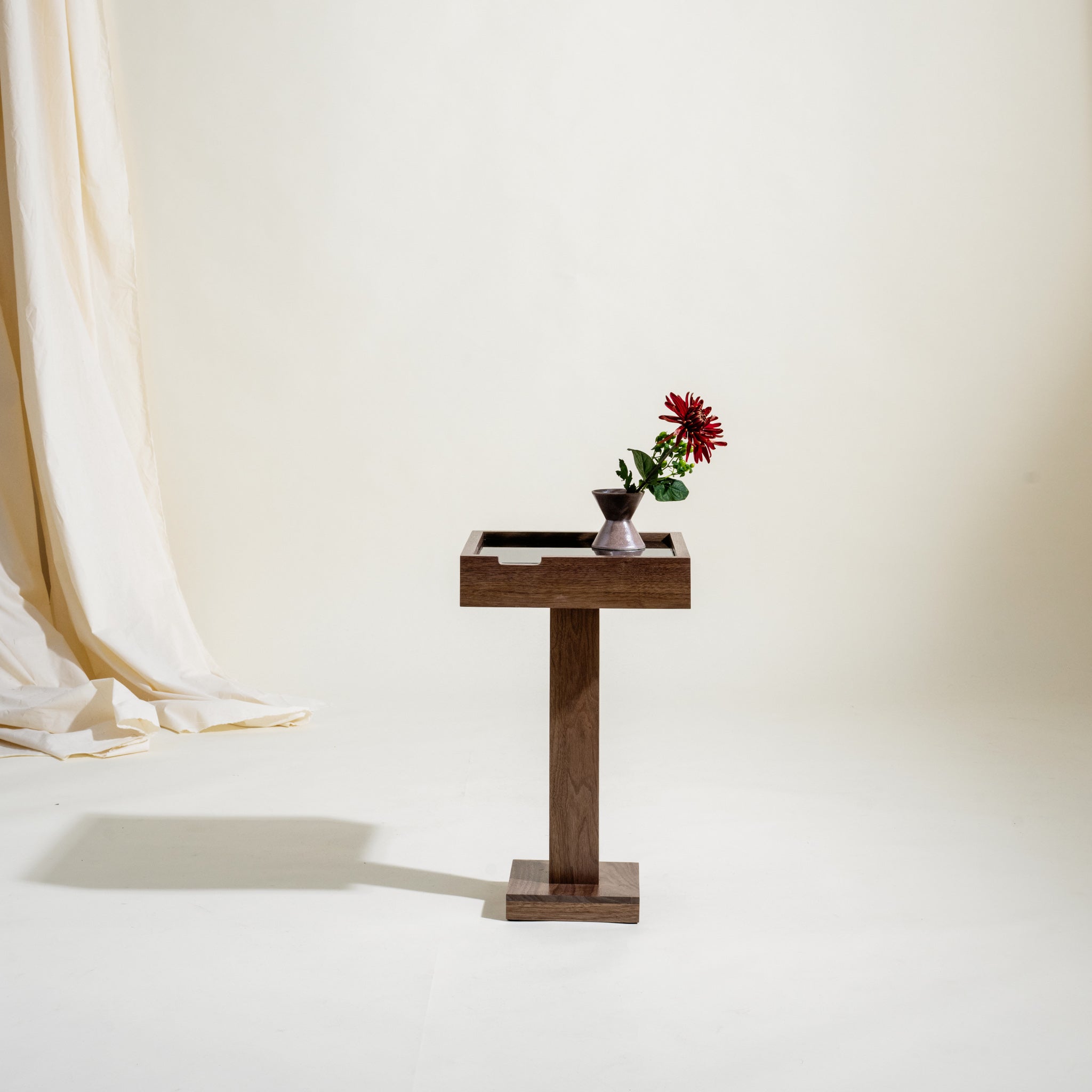 Handcrafted wooden side table with a vase of flowers on a white background. Table has a small square base on the floor and a square column coming up from the base with a rectangular top that is larger is area than the base.